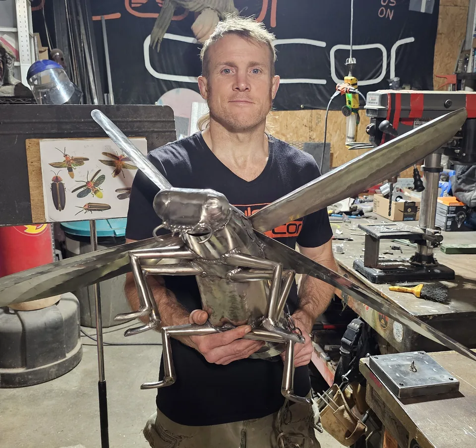 Dustin Weatherby in his SculptDecor studio holding a large welded-steel grasshopper sculpture