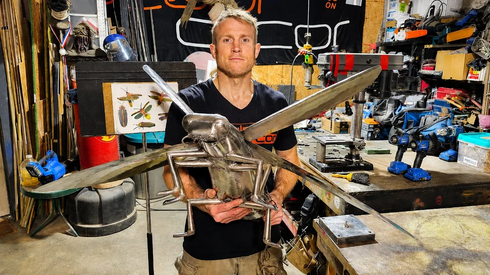 Sculptor Dustin Weatherby in his studio holding a large welded-steel grasshopper, SculptDecor banner behind him