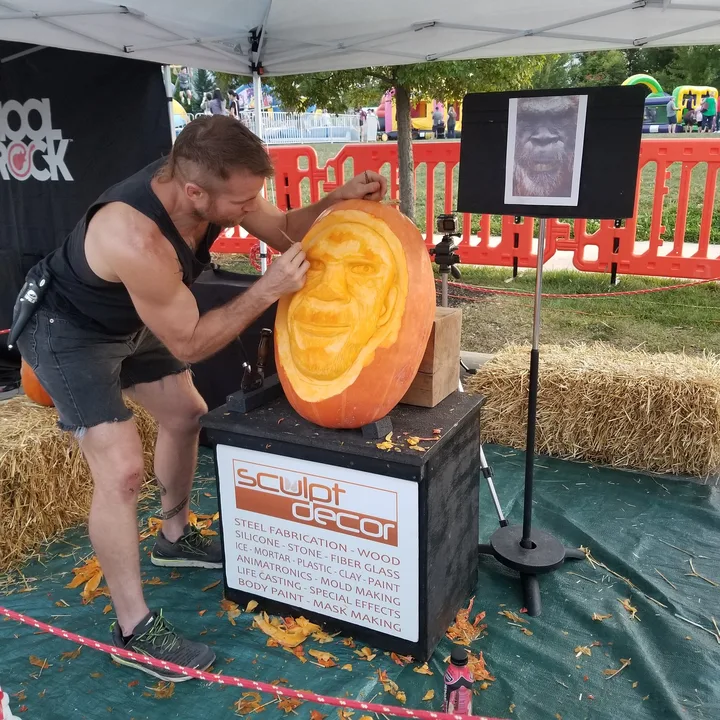 Dustin Weatherby carving a portrait into a giant pumpkin at an outdoor festival beneath a SculptDecor banner